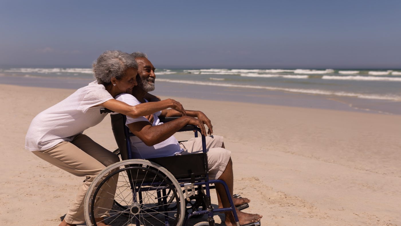 Pareja de adultos mayores en la playa; ella abraza a su esposo, que va en silla de ruedas