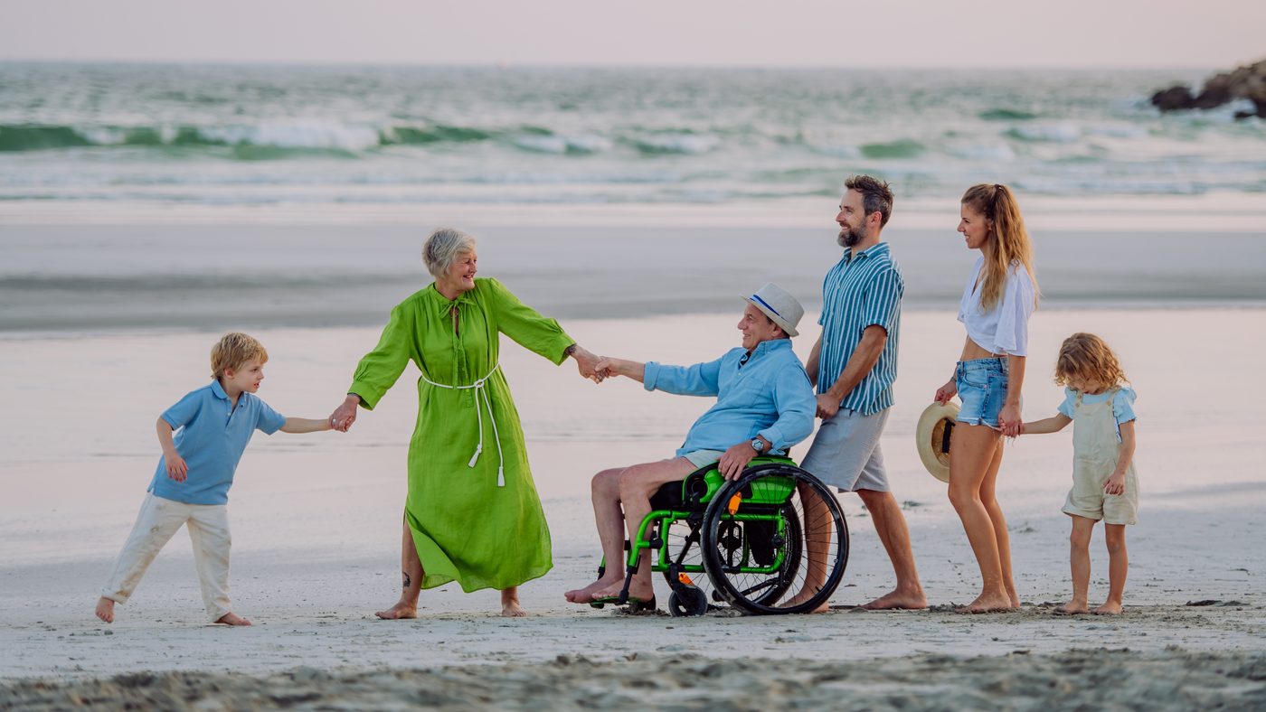 Familia de tres generaciones pasea por la playa con el abuelo en silla de ruedas